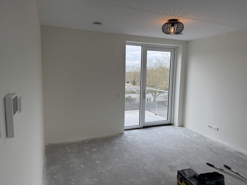 Lege kamer in een nieuw appartement met lichte wanden, een balkondeur en een kale betonvloer