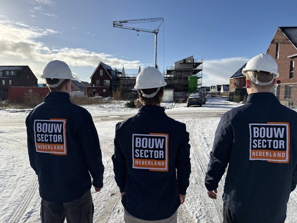 Drie vakmensen van Bouwsector Nederland met witte helm en logo op de rug kijken naar nieuwbouwhuizen in aanbouw met een bouwkraan